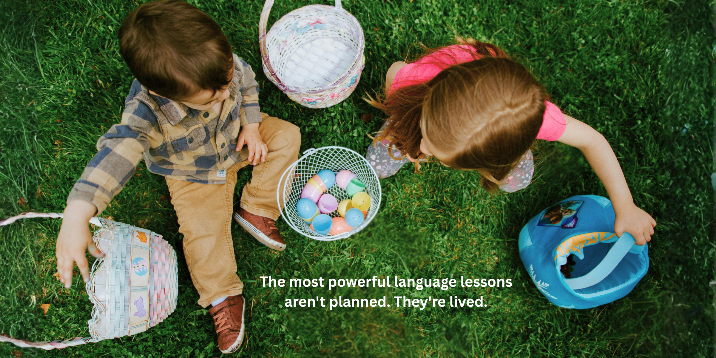 Two children sit on green grass. One child wears a plaid shirt and beige pants, the other a pink top. Between them are two baskets filled with colorful eggs. Another basket with eggs is nearby. At the bottom, the text reads: 'The most powerful language lessons aren't planned. They're lived.'